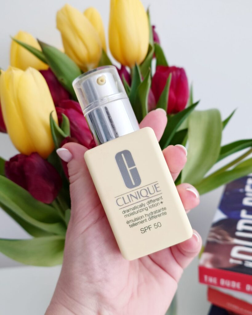 Kobieca dłoń trzymająca żółte opakowanie emulsji nawilżającej Clinique Dramatically Different Moisturizing Lotion+ z filtrem SPF 50. W tle widać świeże, żółte i bordowe tulipany.