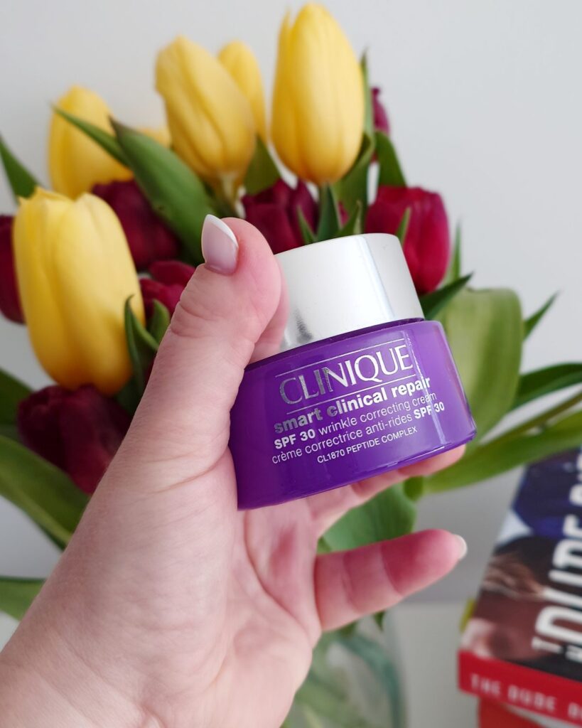 Zbliżenie na fioletowy słoiczek kremu Clinique Smart Clinical Repair SPF 30 trzymany w dłoni na tle bukietu wiosennych tulipanów. Widoczny srebrny korek i napisy na opakowaniu.