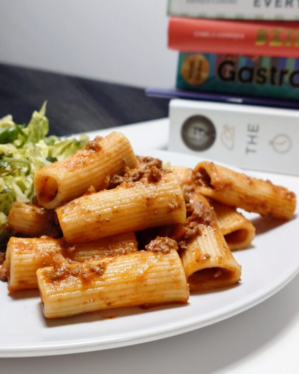 Porcja makaronu rigatoni z sosem bolognese i zieloną sałatą na białym talerzu, w tle stos książek kulinarnych.