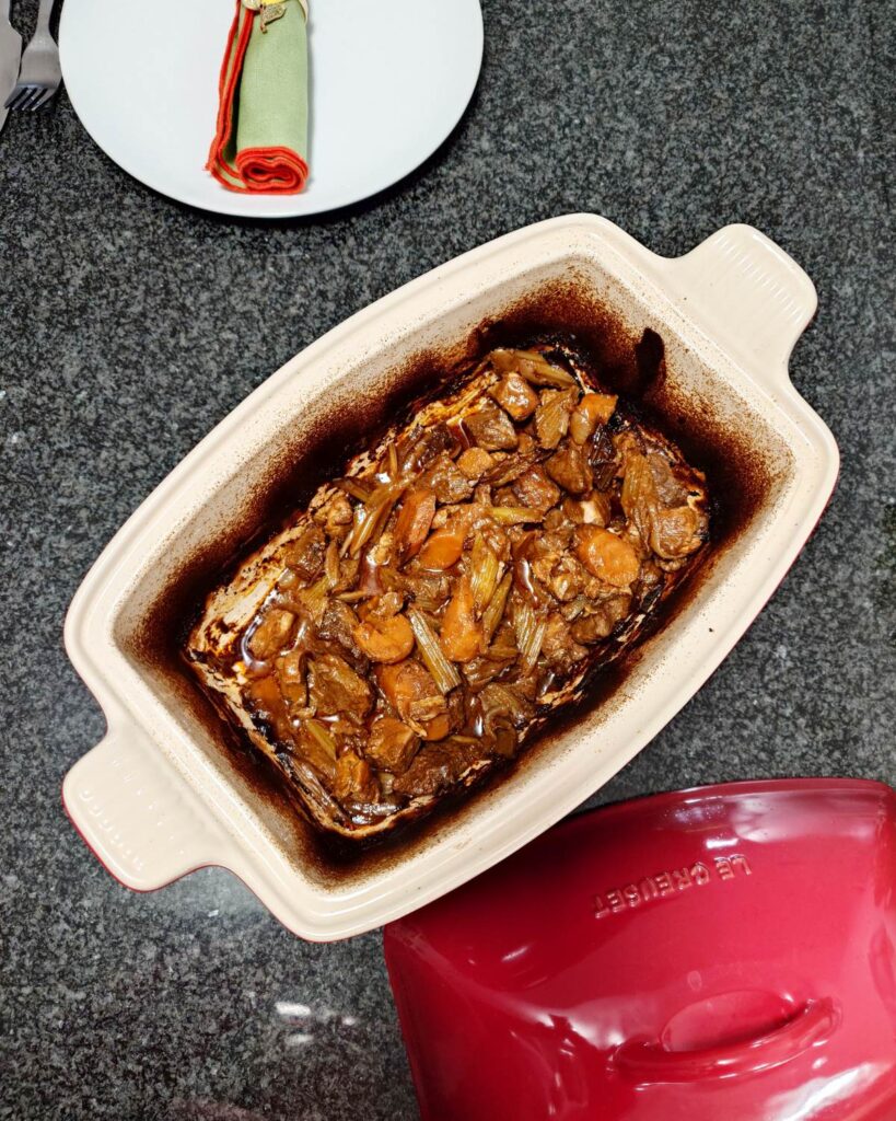 Prostokątne naczynie do zapiekania Le Creuset wypełnione ciemnym gulaszem wołowym w piwie Guinness, marchewką i selerem naciowym. Obok naczynia leży czerwona pokrywka Le Creuset, a na białym talerzu widać zieloną serwetkę z metalowym listkiem.