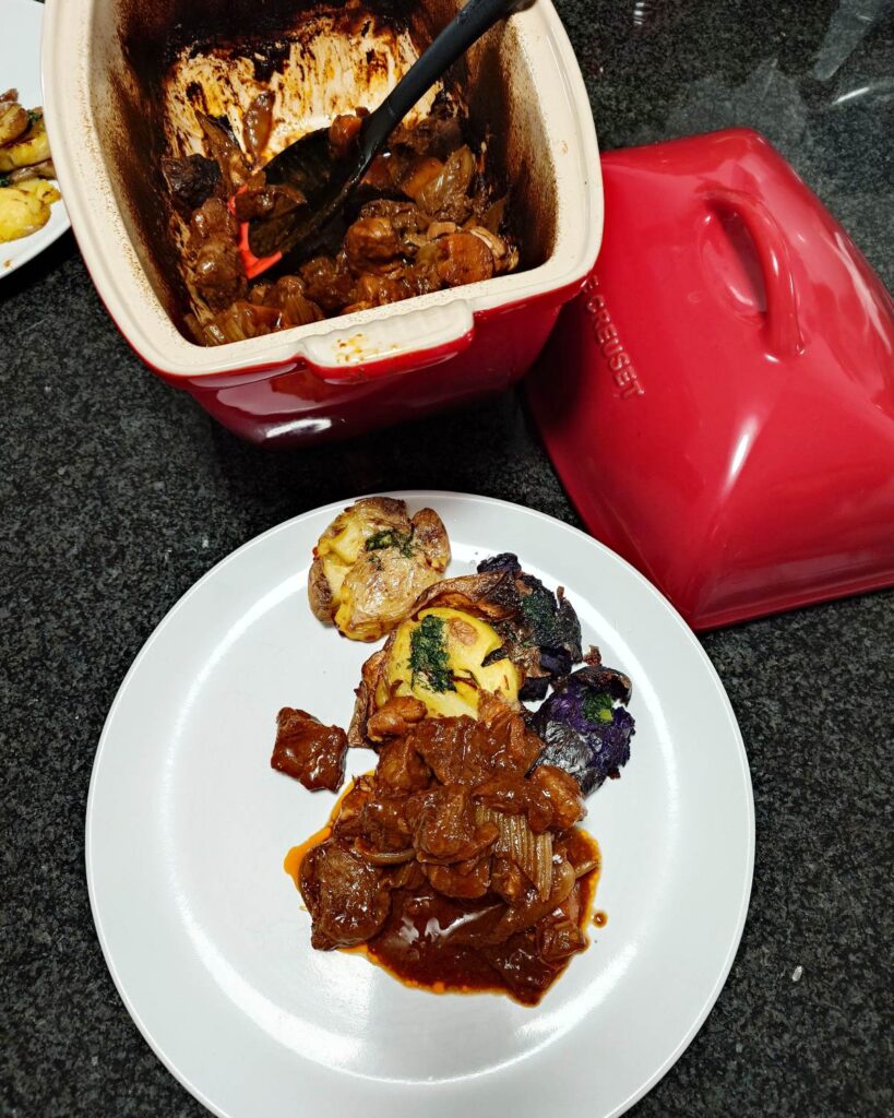 Porcja gulaszu wołowego w Guinnessie podana na białym talerzu obok pieczonych ziemniaczków smashed w dwóch kolorach (fioletowe i zwykłe), polanych masłem ziołowym. W rozmytym tle widać czerwone naczynie Le Creuset.