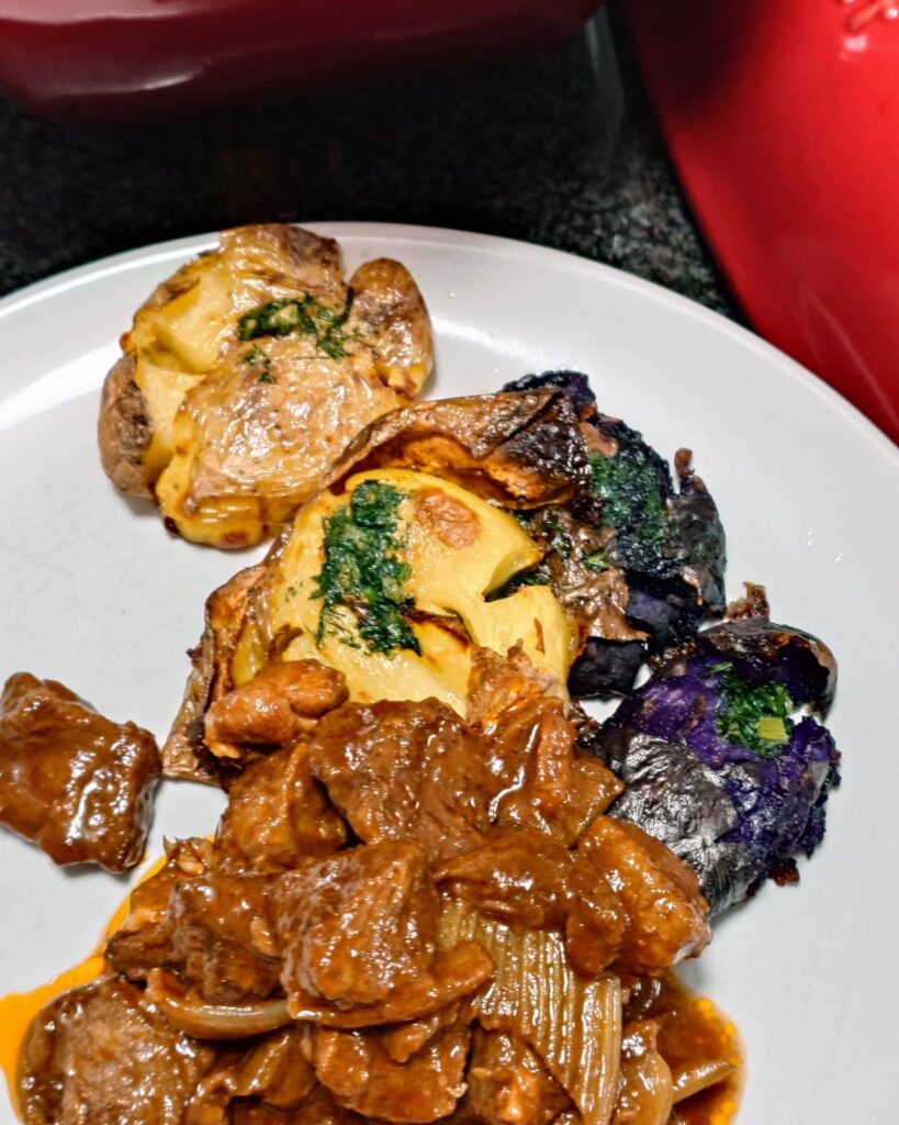 Zbliżenie na talerz z gulaszem wołowym w piwie Guinness. Na pierwszym planie chrupiące fioletowe i zwykłe ziemniaczki smashed z roztapiającym się masłem ziołowym, obok kawałek miękkiej wołowiny w gęstym, ciemnym sosie.