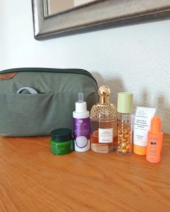 Otwarta oliwkowa kosmetyczka podróżna stojąca na drewnianym blacie, obok ułożone kosmetyki premium: perfumy Guerlain Aqua Allegoria, serum BasicLab i kremy w opakowaniach podróżnych.