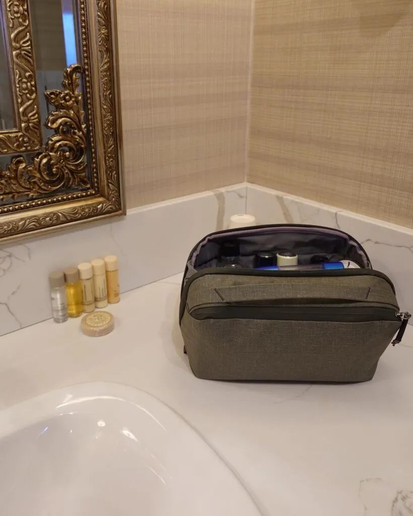 Zamknięta zielona kosmetyczka podróżna Peak Design stojąca na marmurowym blacie w eleganckiej łazience obok ozdobnego, złotego lustra i hotelowych miniatur.
