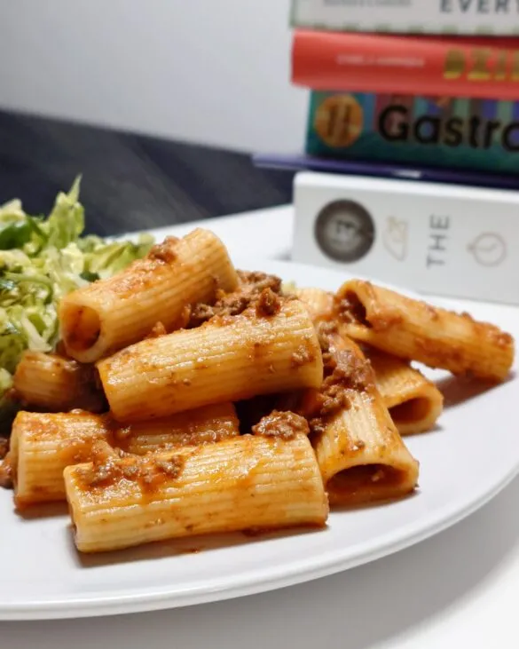 Porcja makaronu rigatoni z sosem bolognese i zieloną sałatą na białym talerzu, w tle stos książek kulinarnych.