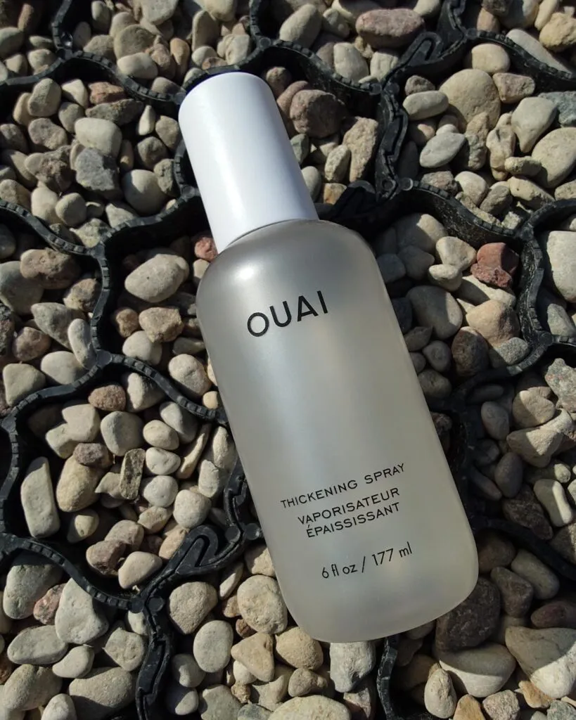 Butelka sprayu pogrubiającego Ouai Thickening Spray na jasnych kamieniach. Zdjęcie produktowe kosmetyku do objętości włosów w słońcu.